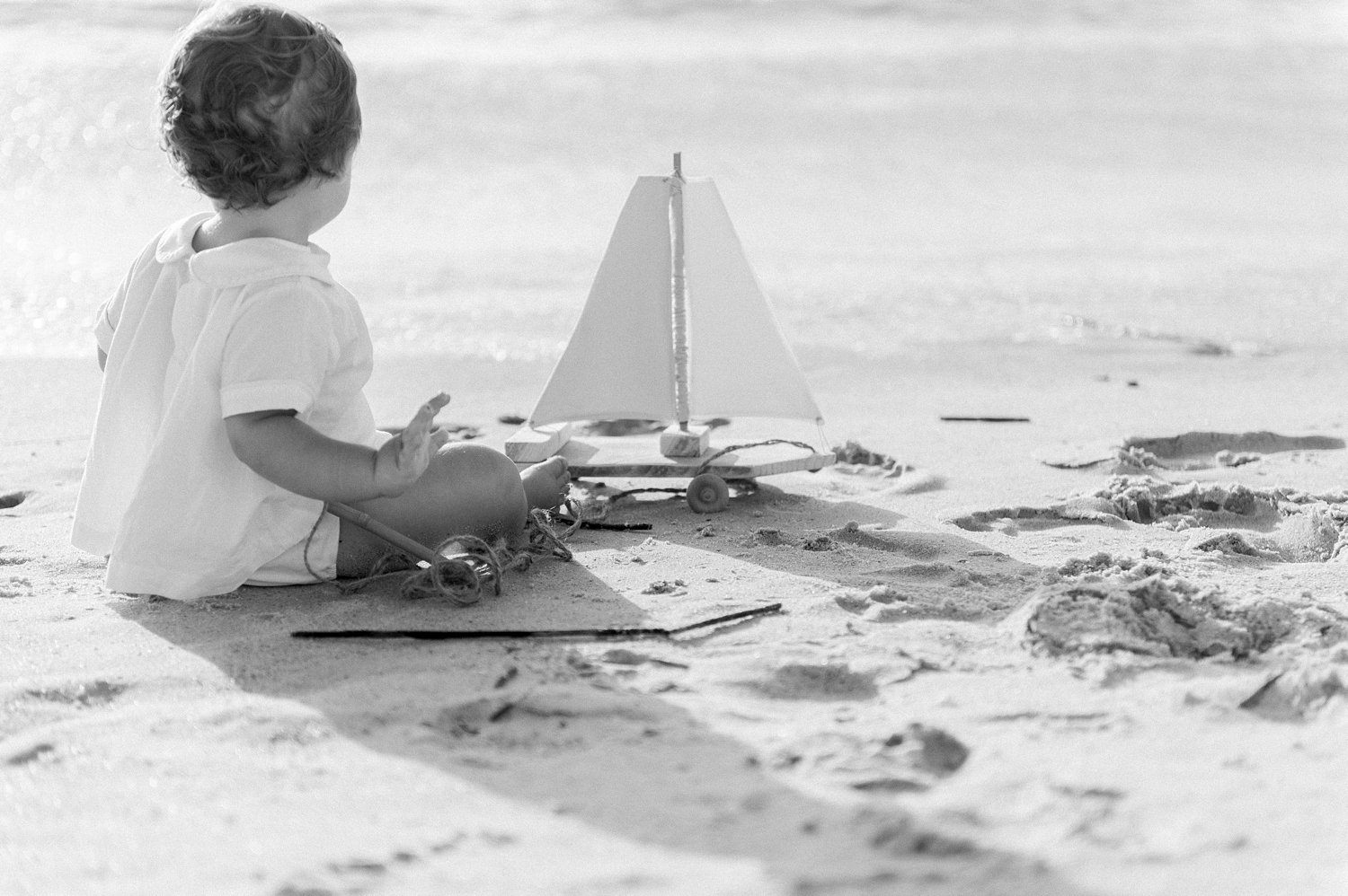 black and white portrait of a one year old little boy with a boat pull toy on the beach