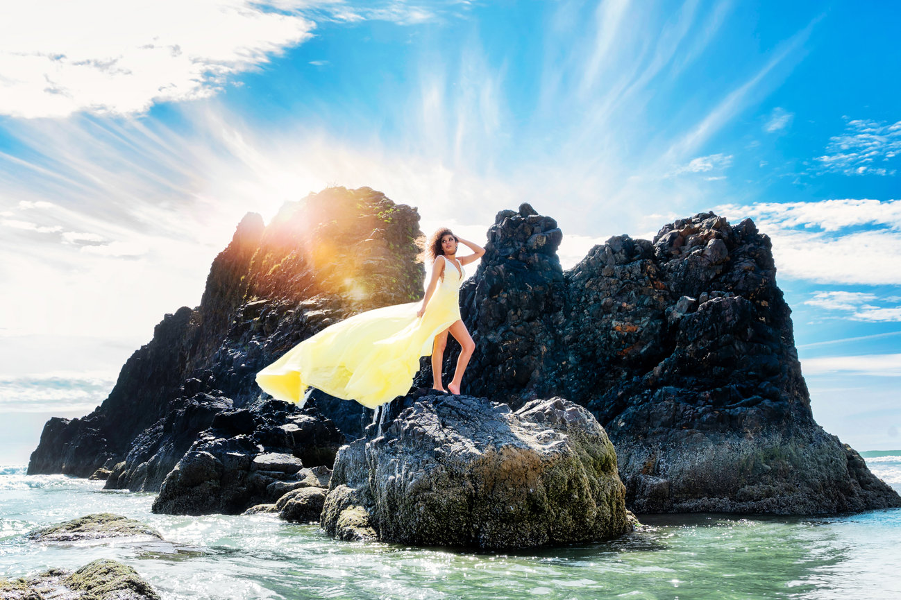 Woman in a yellow dress poses on a rocky beach against a bright sky with sun rays.