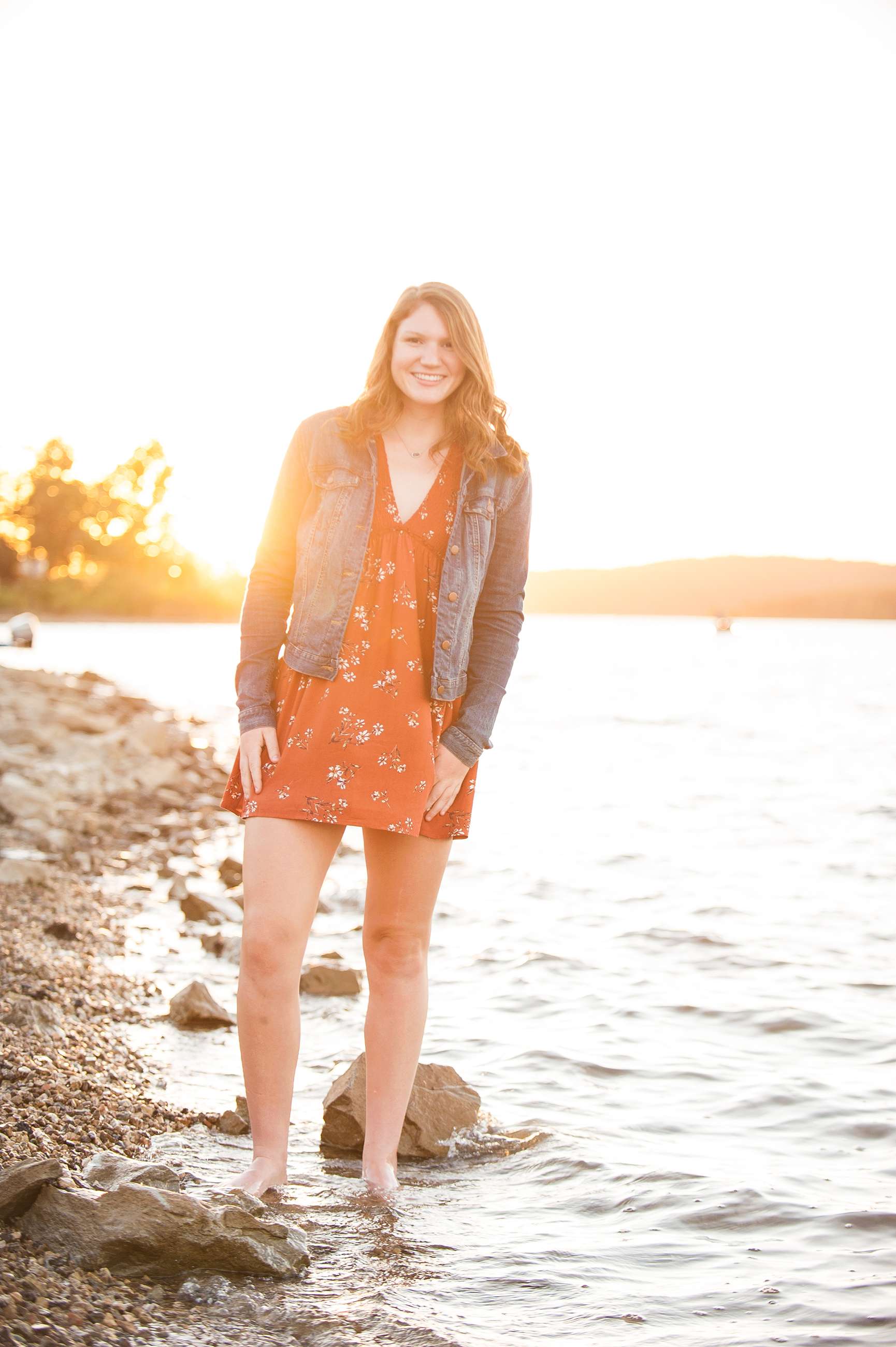 senior girl in jean jacket and orange dress at sunset standing on shore of Table Rock Lake posing for portrait