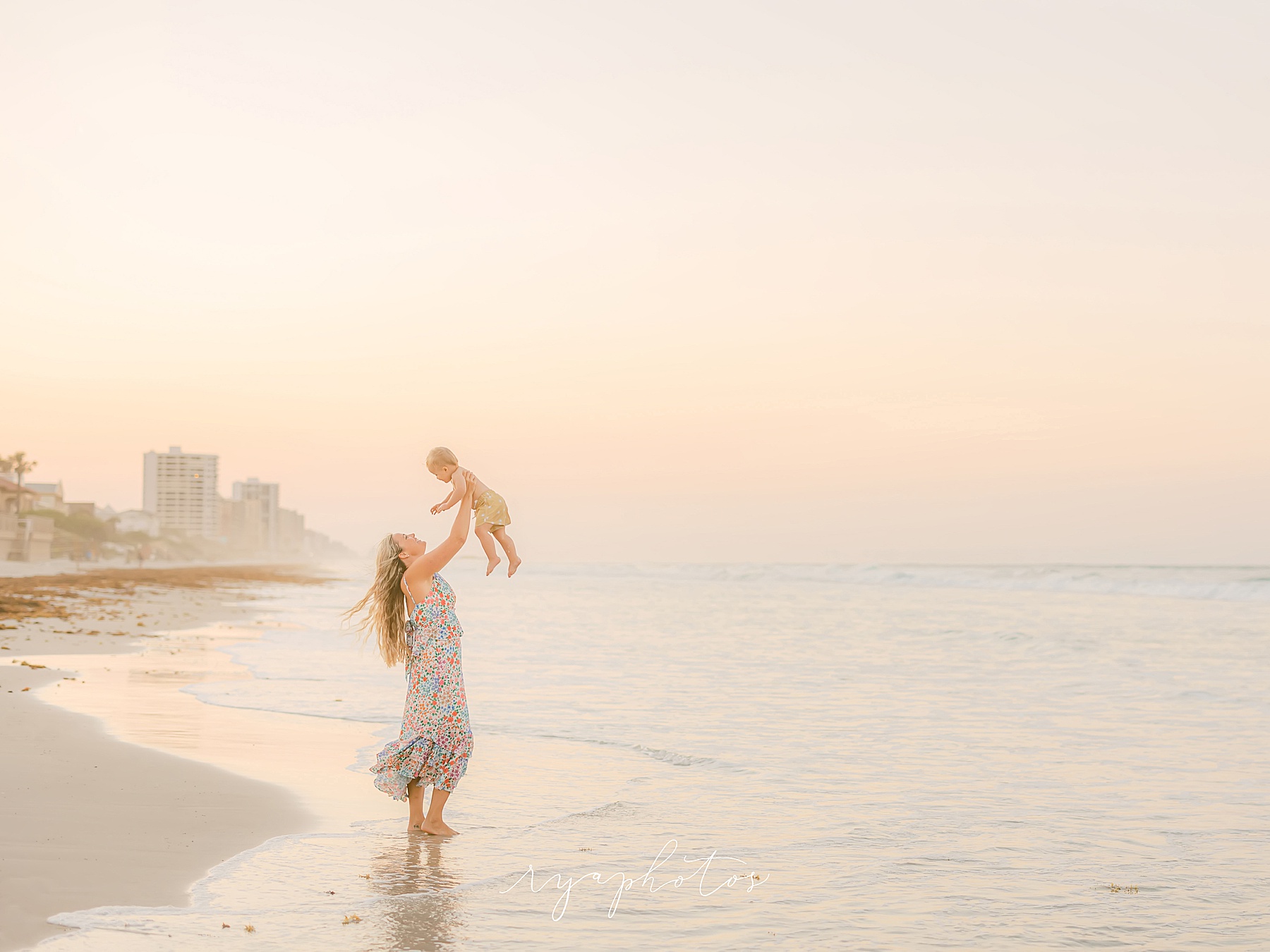 Woman holding baby at sunset on beach in pastel colored dress dreamy