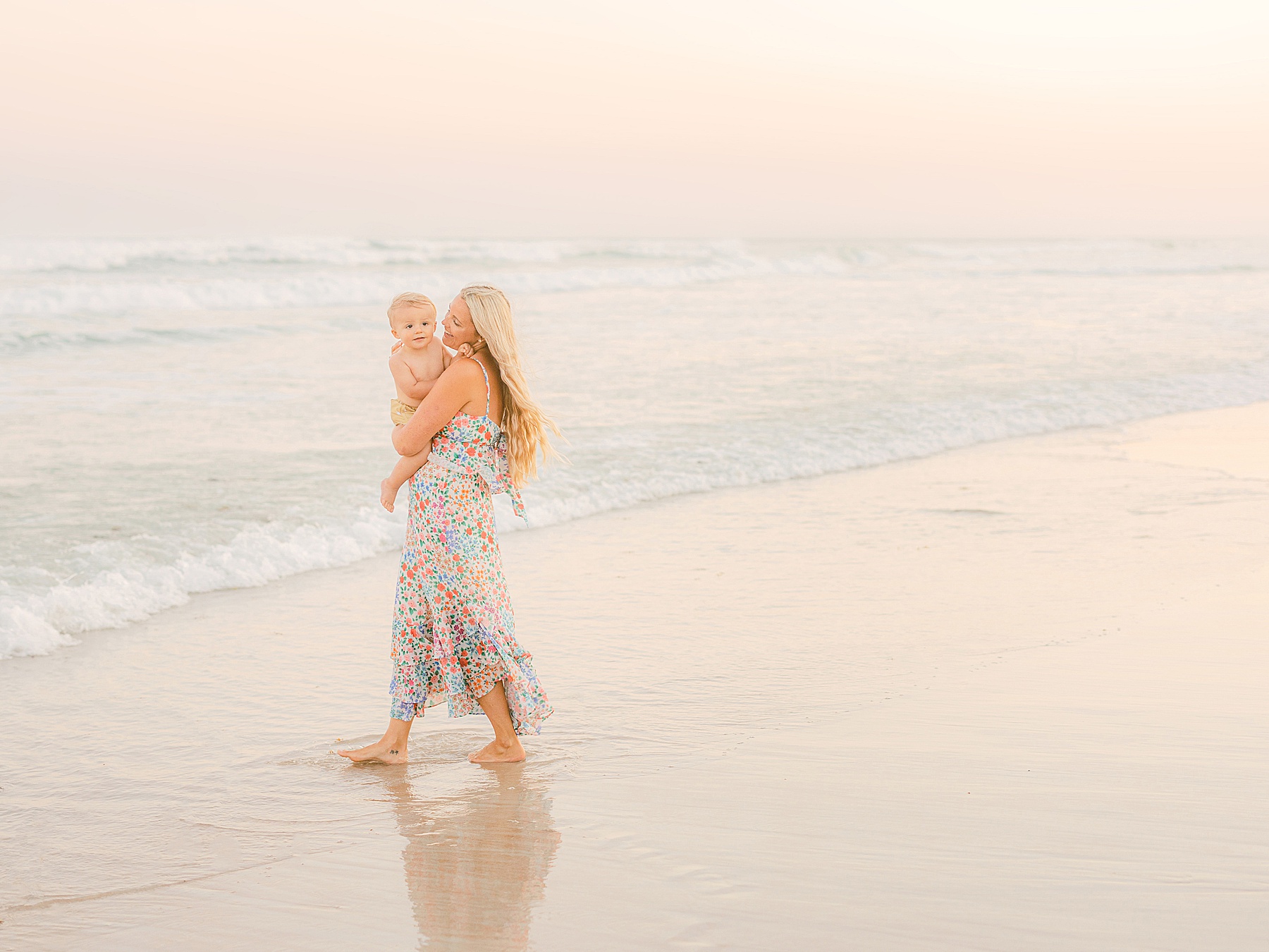 mom holding baby boy in the air on the beach in a pastel sky
