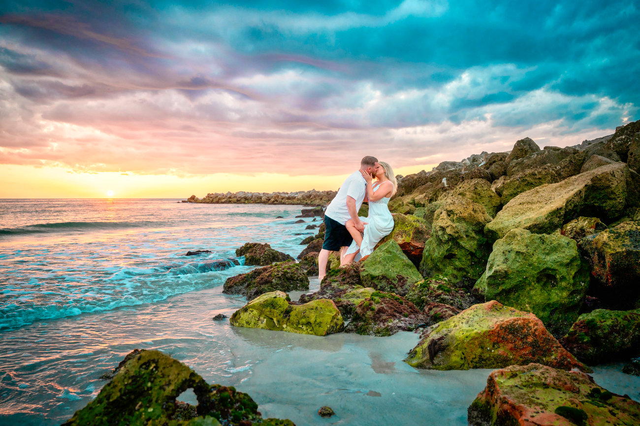 Couple kissing on rocky beach at sunset, with colorful skies and gentle waves in the background.