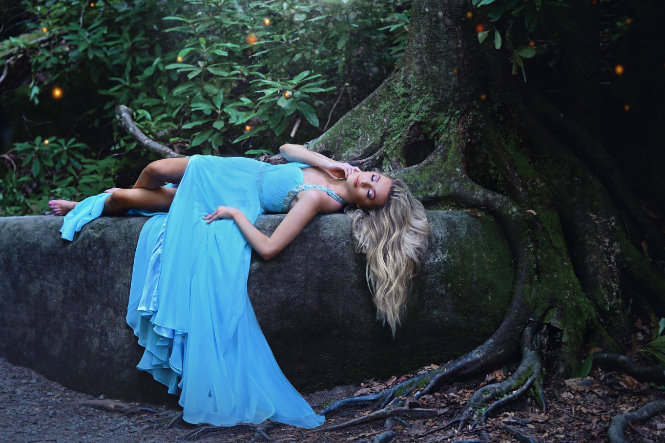 A woman in a flowing blue dress reclines on a moss-covered rock, surrounded by lush greenery.