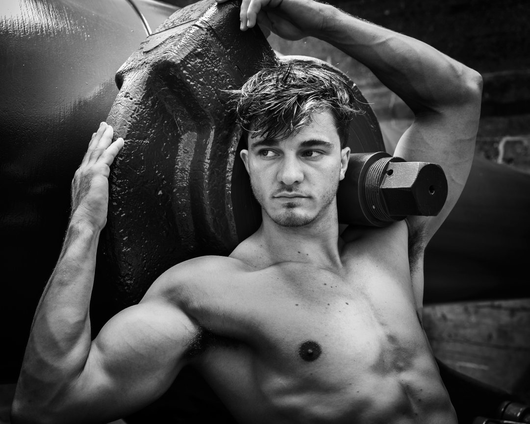 Muscular man posing shirtless with a large weight on his shoulder, in a dramatic black and white setting.