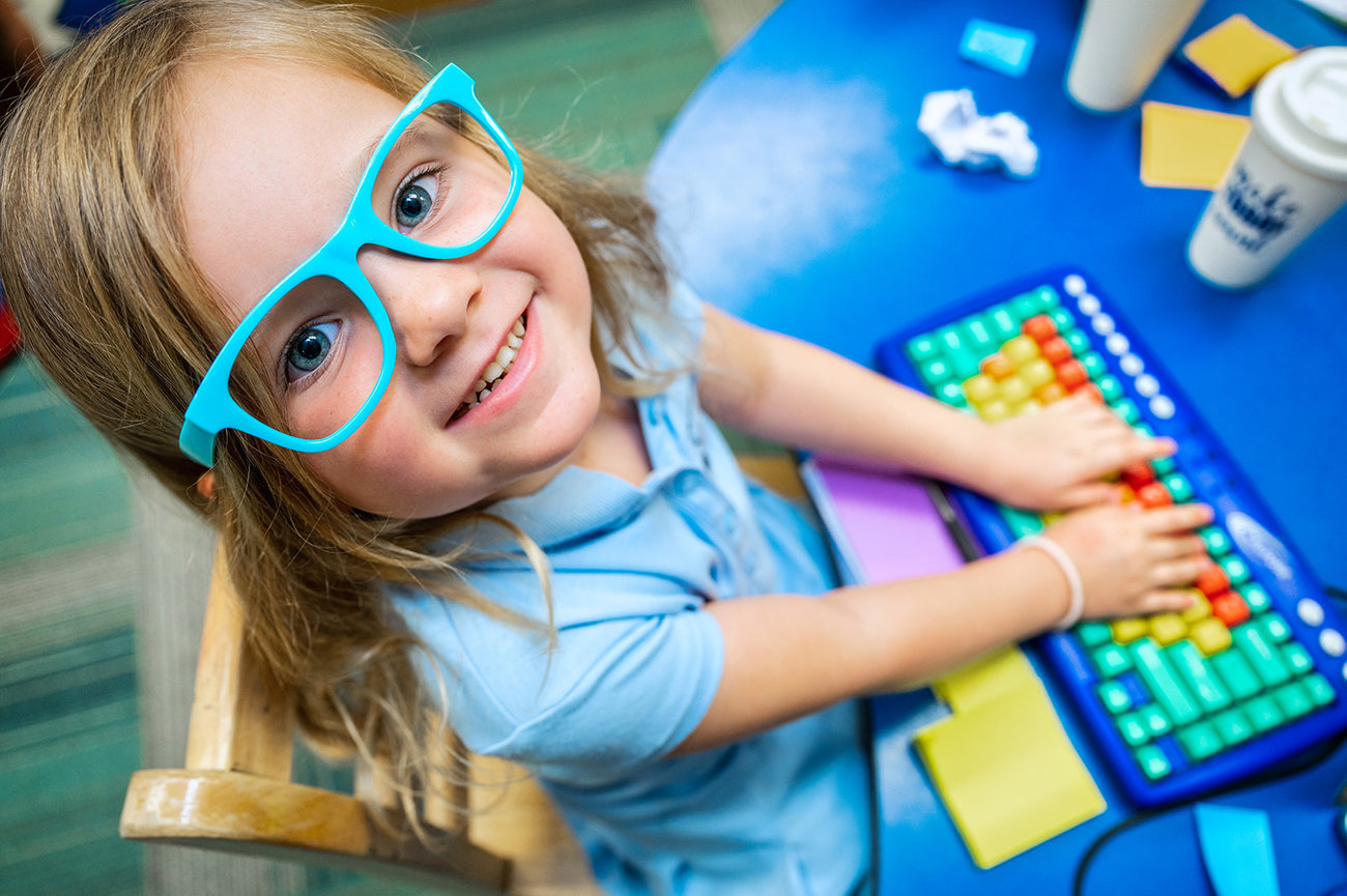 Smiling child with glasses typing on a colorful keyboard at a blue table, surrounded by sticky notes.