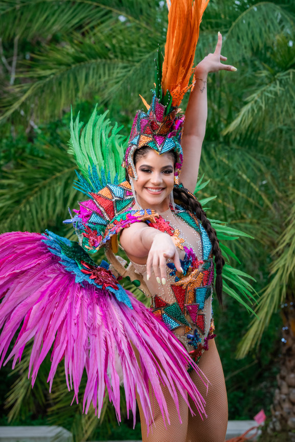 Colorful dancer in a vibrant feather costume poses joyfully against a tropical backdrop.