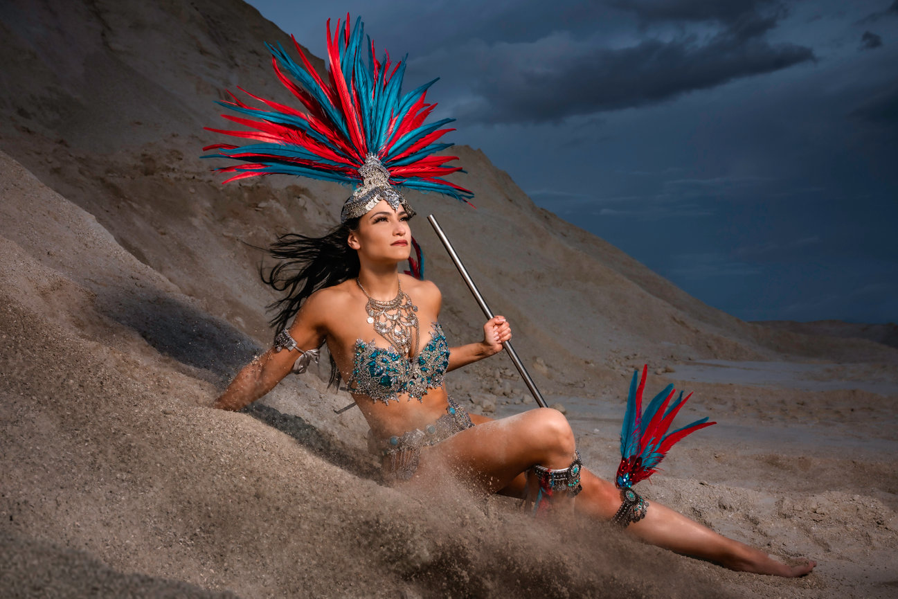 Model in vibrant costume with feathers poses in sandy landscape, showcasing a dramatic look against a moody sky.