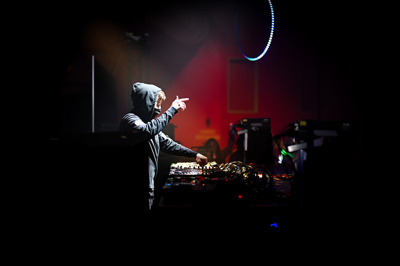 DJ performing live with a hooded sweatshirt, focused on mixing music at a dimly lit venue.