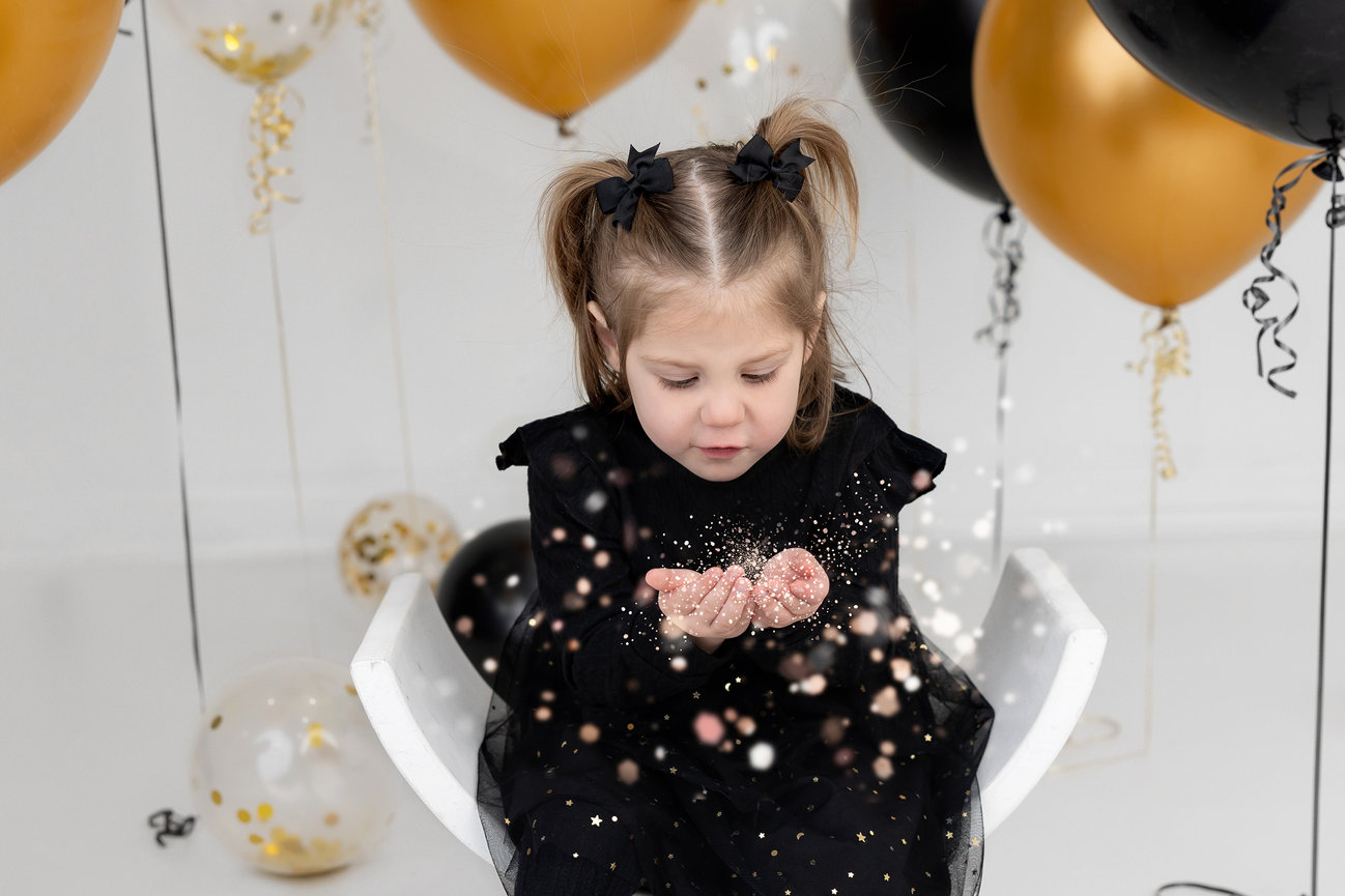 Child in black dress sitting, holding confetti with gold and black balloons in the background.