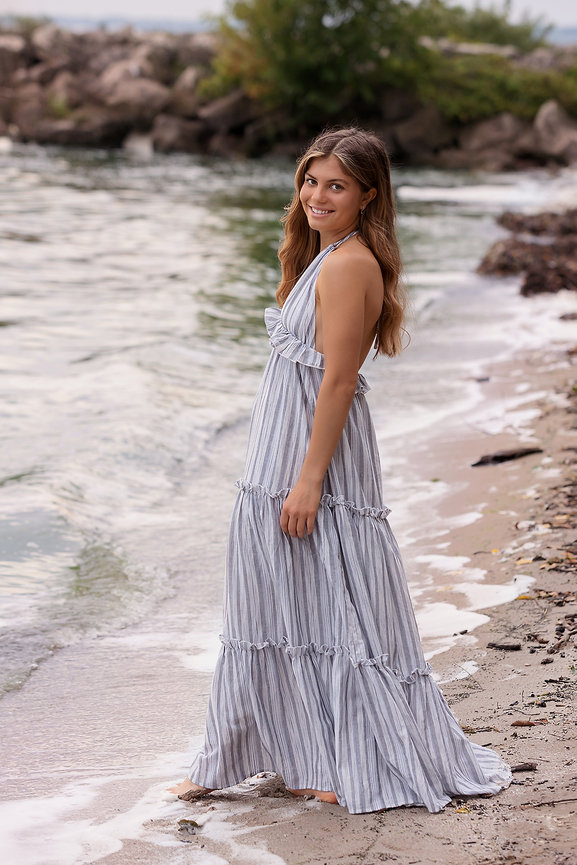 Woman in a striped dress walking along a sandy beach with rocks and water in the background.