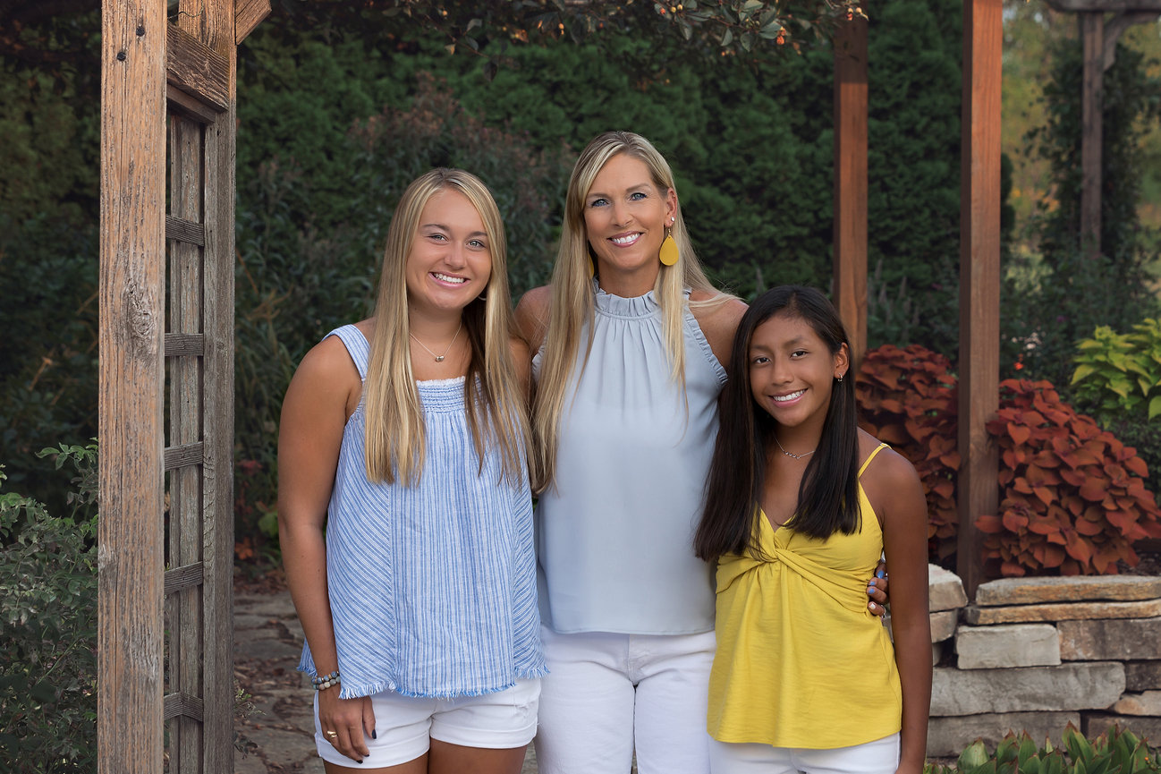 Three people posing outdoors in a garden setting, with wooden structures and greenery in the background.