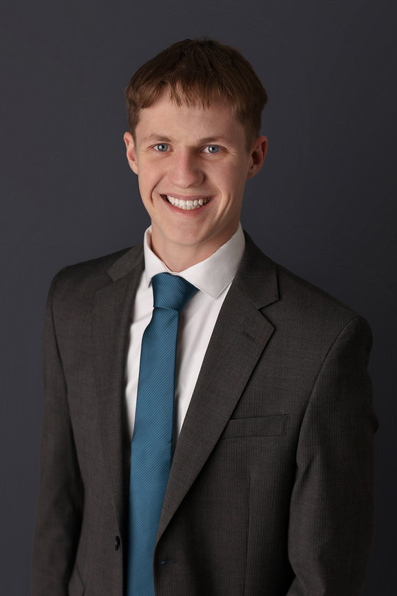 Smiling person in a suit with a blue tie, standing against a dark background.