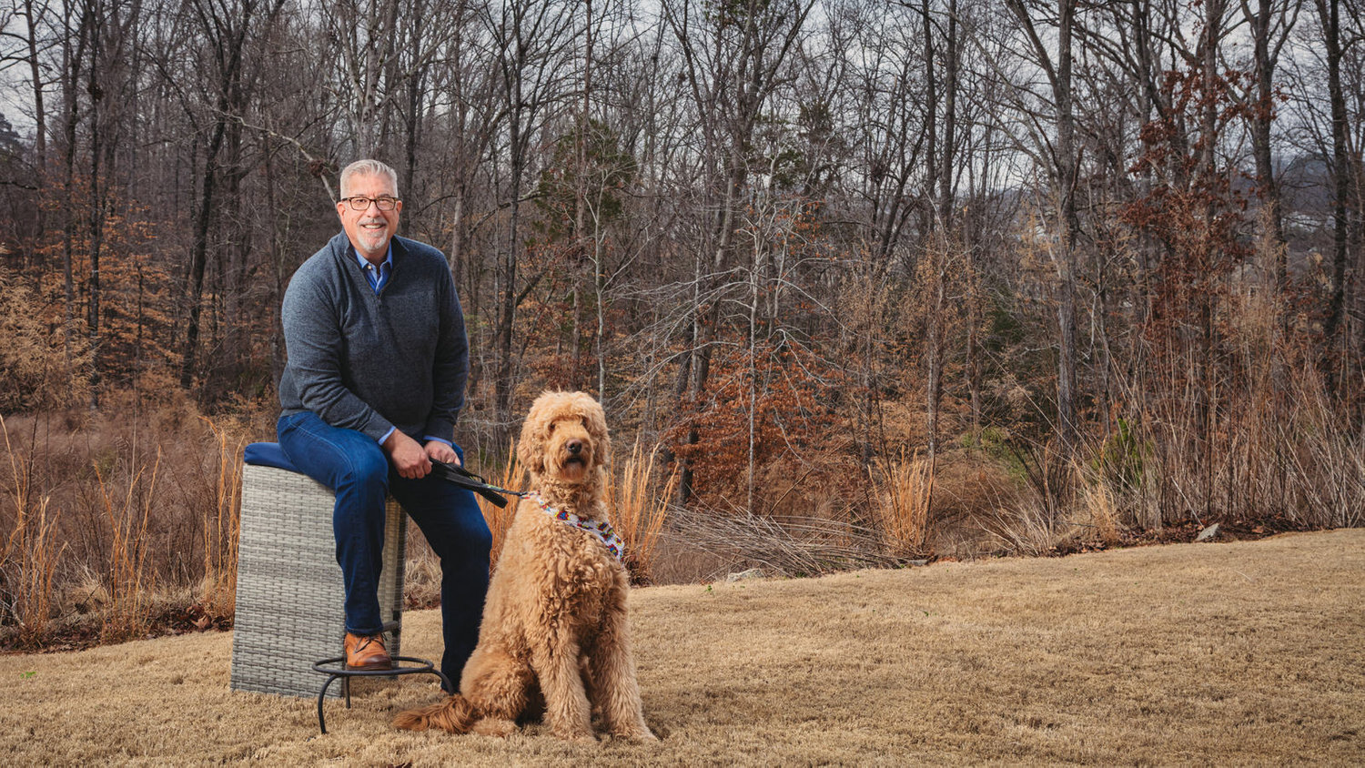Man sitting on a wicker stool in a field, holding a leash attached to a large, fluffy dog. Bare trees in the background.