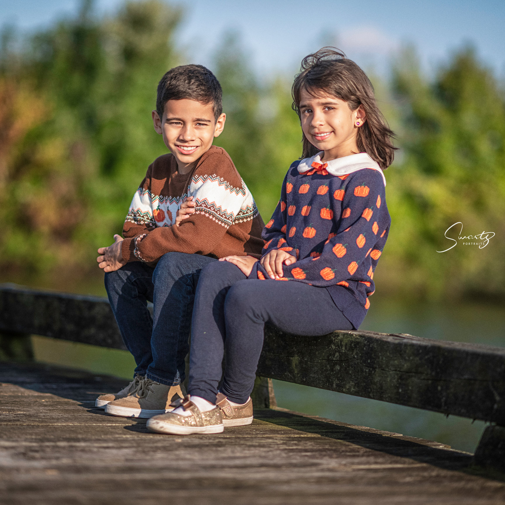 Two young children, boy and a girl, sit on a wood edge next to water.