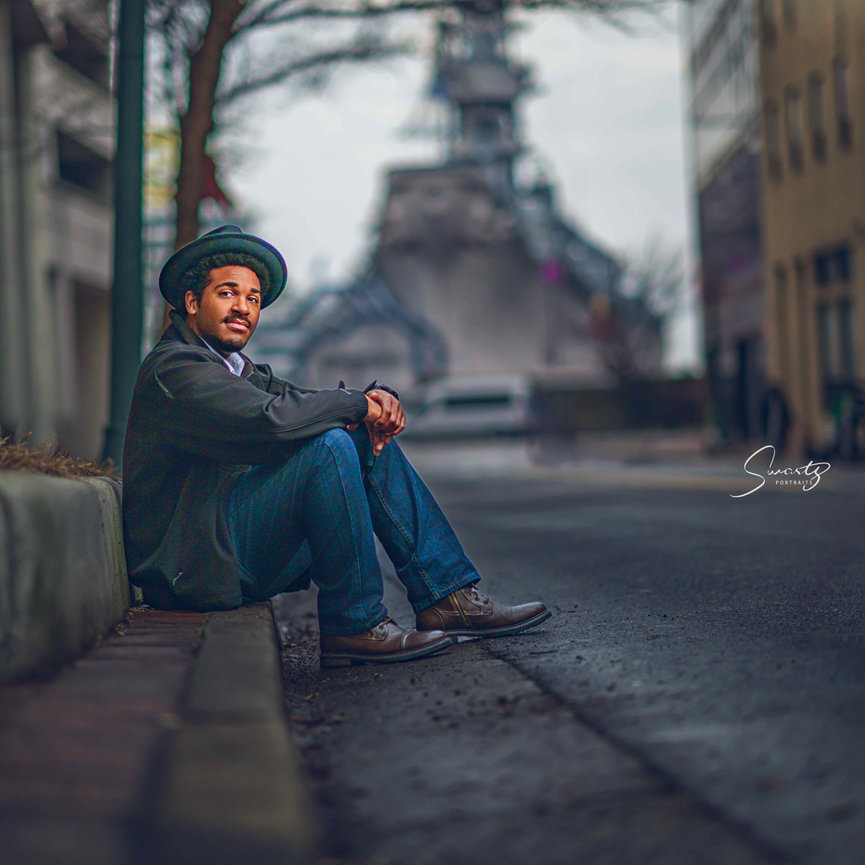 Senior boy in fedora and dark blazer seated on urban sidewalk curb with deep architectural background — fine art editorial environmental senior portrait by Master Photographer Ty Swartz, Swartz Portraits Houston Texas inner-loop senior photographer