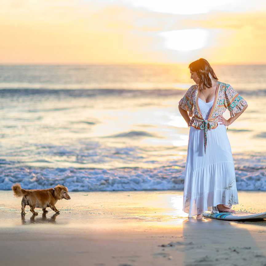 Beach sunrise senior portrait in Galveston, girl in white peasant dress with golden first light over Gulf horizon, dachshund dog walking in surf, cinematic fine art beach session by Ty Swartz, Swartz Portraits Spring Texas senior beach photographer