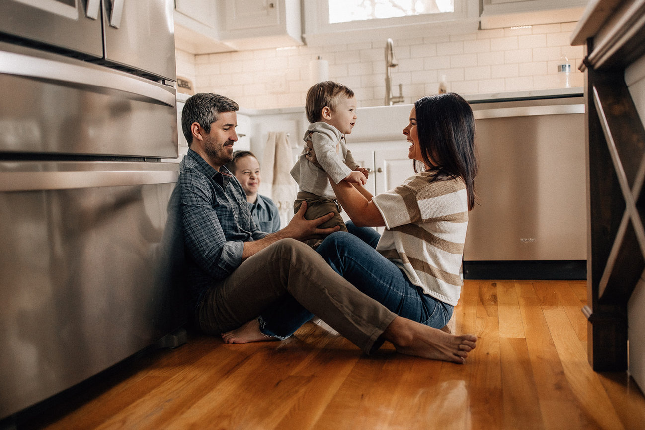 A family of four sit together on a hardwood floor in a kitchen during a Huntsville Alabama family photoshoot.