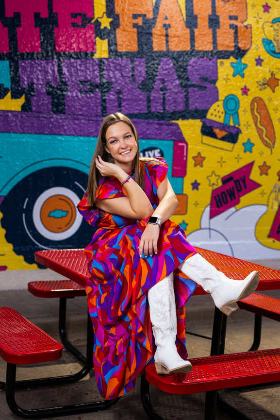 Beautiful high school senior girl in colorful red dress with white boots sitting on a red picnic table in font of a Texas State Fair mural.