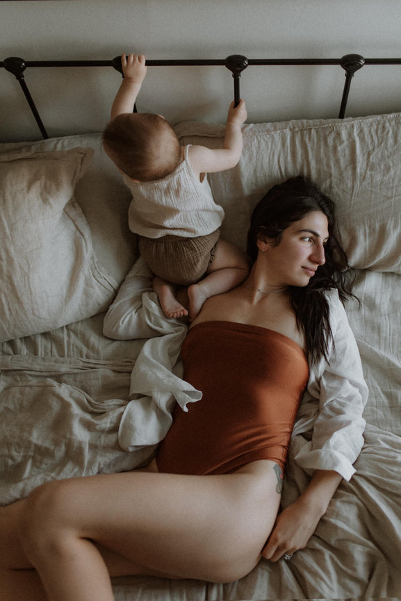 A mother lying on a bed and gazing out the window while holding her baby in Jersey City, New Jersey.