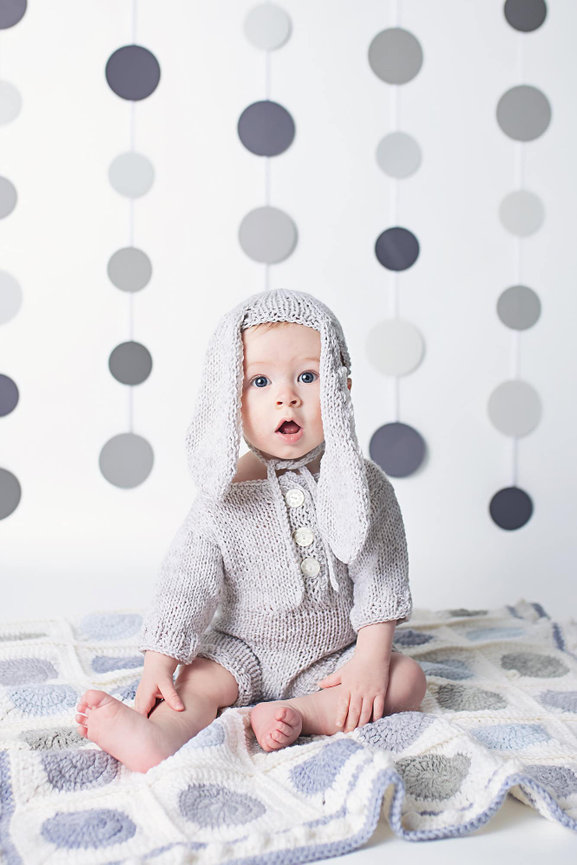A six month old boy sits on a grey and white granny square blanket. He wears a grey romper and a bunny ear bonnet. There are grey circles on the wall behind him.