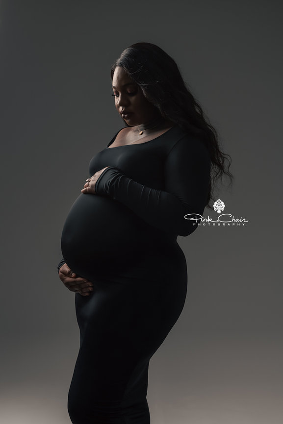 Pregnant woman in a black dress holding her belly against a dark background, looking down.