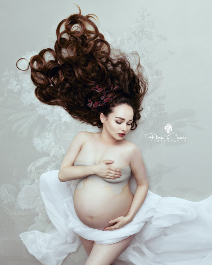 Pregnant woman with long flowing hair, covering chest, draped in white fabric, lying on a floral-patterned backdrop.