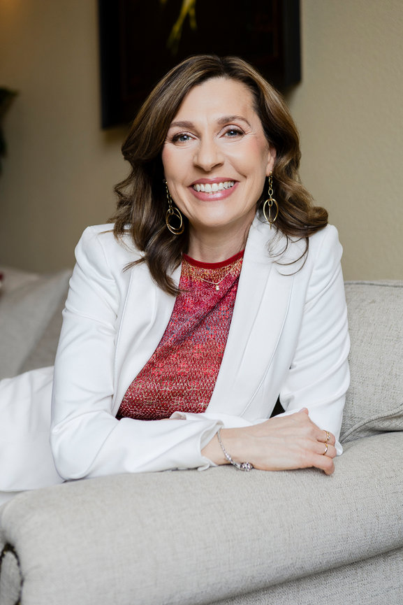 Woman smiling while leaning on a sofa wearing a white blazer and red top in a warmly lit room.