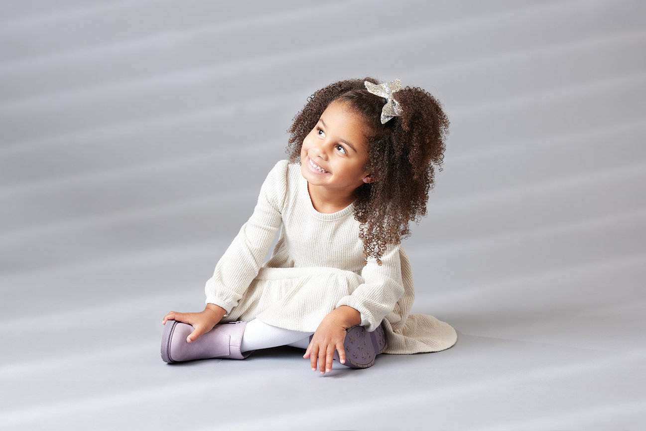 Full-body indoor portrait of a young child sitting on a gray background, softly lit to create a natural light feel with a relaxed and expressive moment