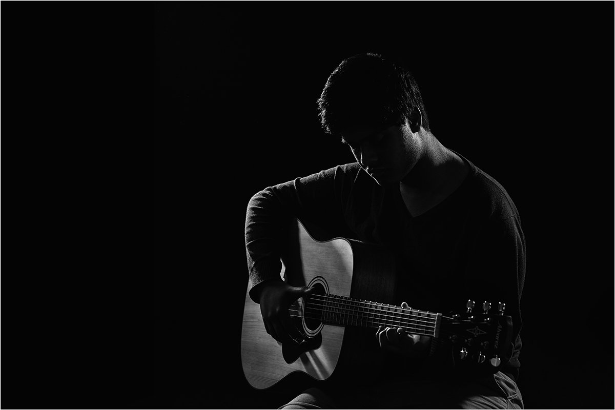 Artistic indoor portrait of a high school senior playing guitar, captured with dramatic low-key lighting to create a sculpted silhouette and a quiet, expressive mood