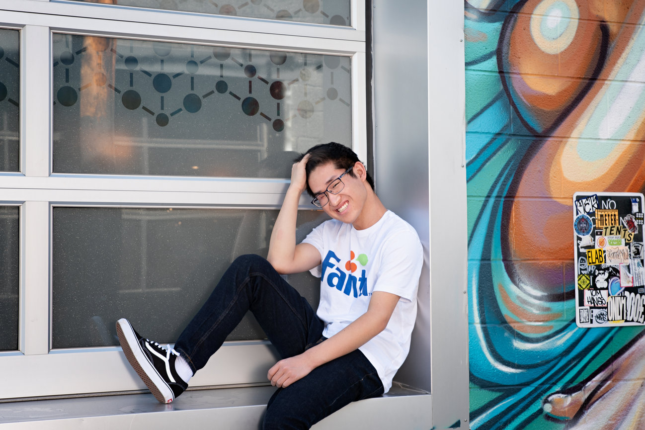 Teen in glasses sits on a window ledge, wearing a Fanta shirt, next to a colorful graffiti wall.