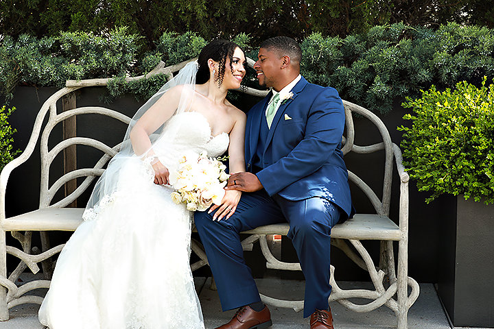 Bride and groom sitting on a decorative bench, smiling at each other, with greenery in the background.