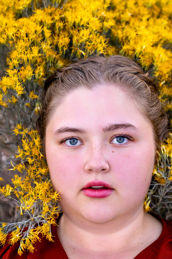 Person with braided hair lying on yellow flowers, looking at the camera, wearing a red top.