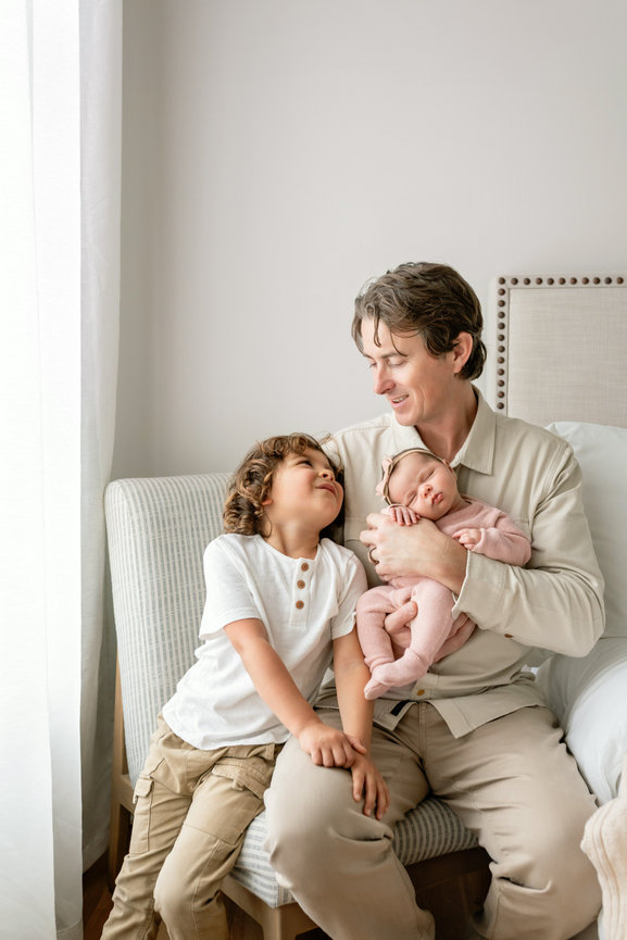 Father holding newborn baby with young child sitting beside him on a bed