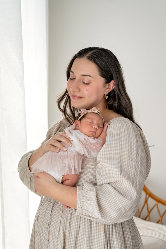 Mother cradling a sleeping newborn baby in a soft pink dress in a bright bedroom