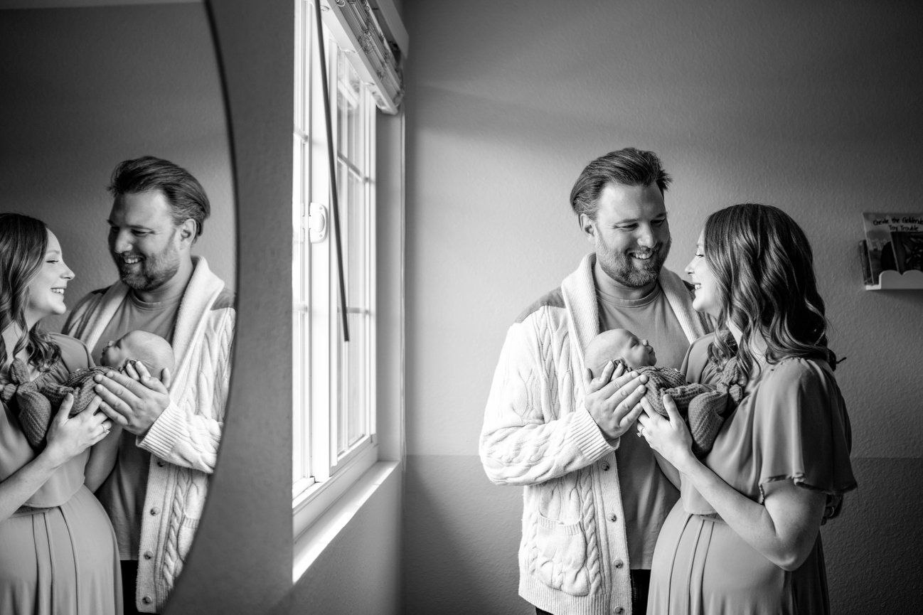 Black and white photo of a smiling couple holding a sleeping newborn by a window