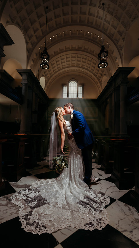 Bride and groom kiss in an elegant, dimly lit church with arched ceiling and decorative hanging lights.