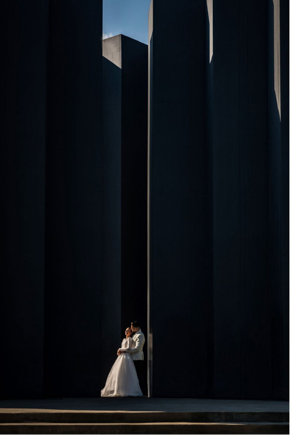 A couple embraces in wedding attire between towering dark architectural columns.