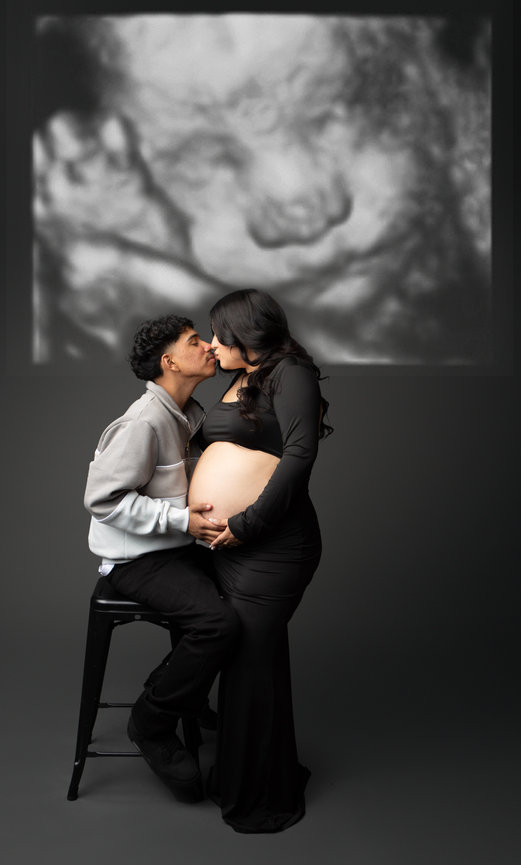 Pregnant couple kissing; woman in black dress, man seated. Ultrasound image in background.