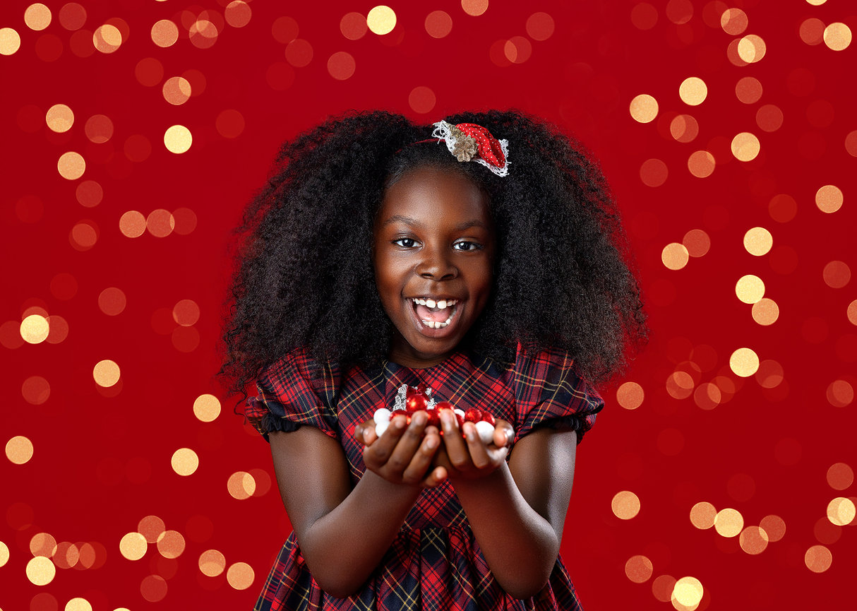 A smiling young girl holding red holiday ornaments against an elegant red Christmas studio backdrop with golden twinkle bokeh lights in Wooster, Ohio.