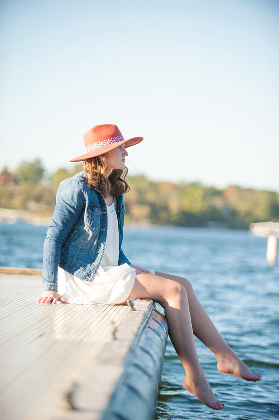 senior girl sitting on dock at Table Rock Lake wearing white dress jean jacket and hat posing for senior portrait