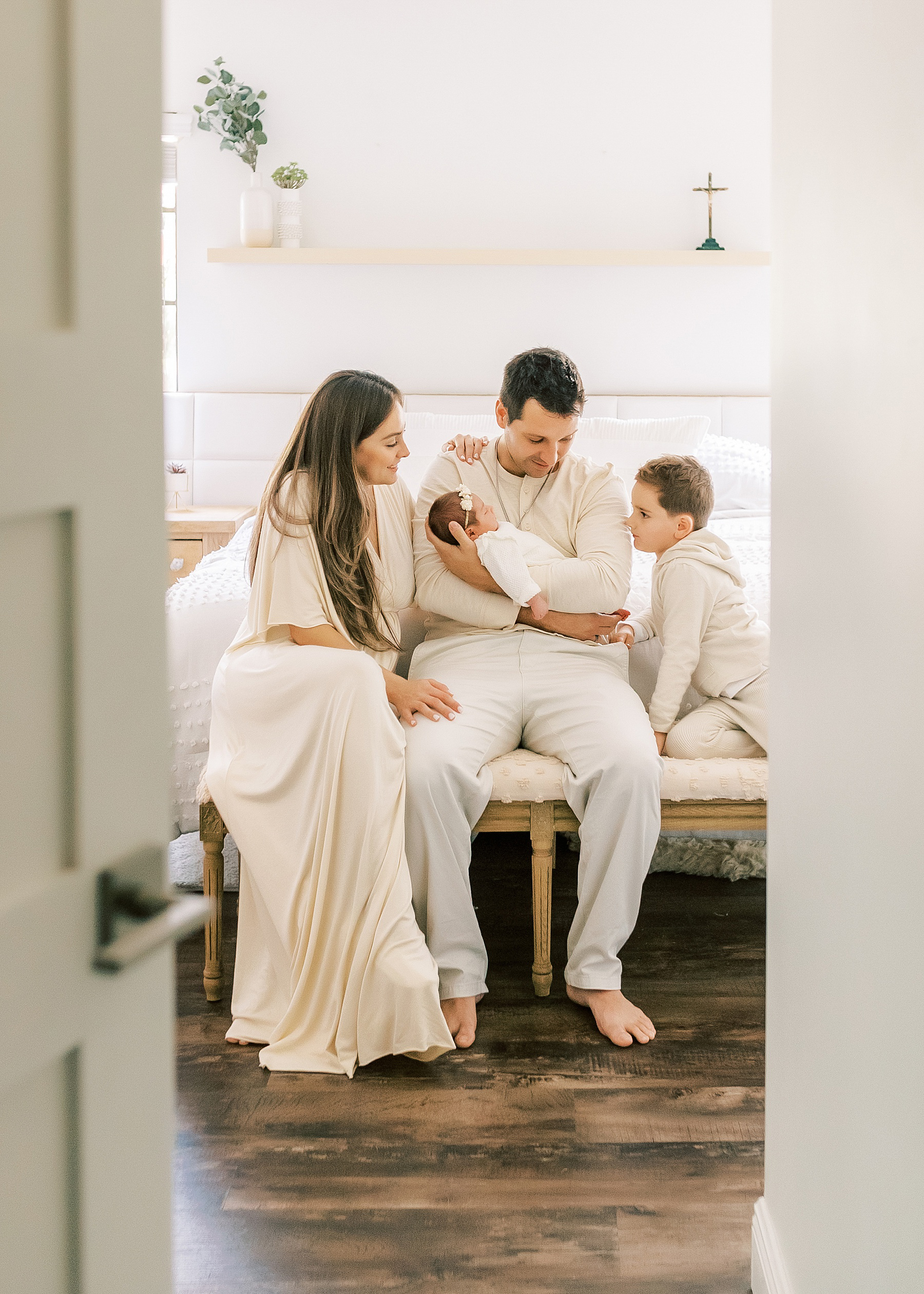 family dressed in tans and creams sitting on bed with white bedding holding newborn baby girl
