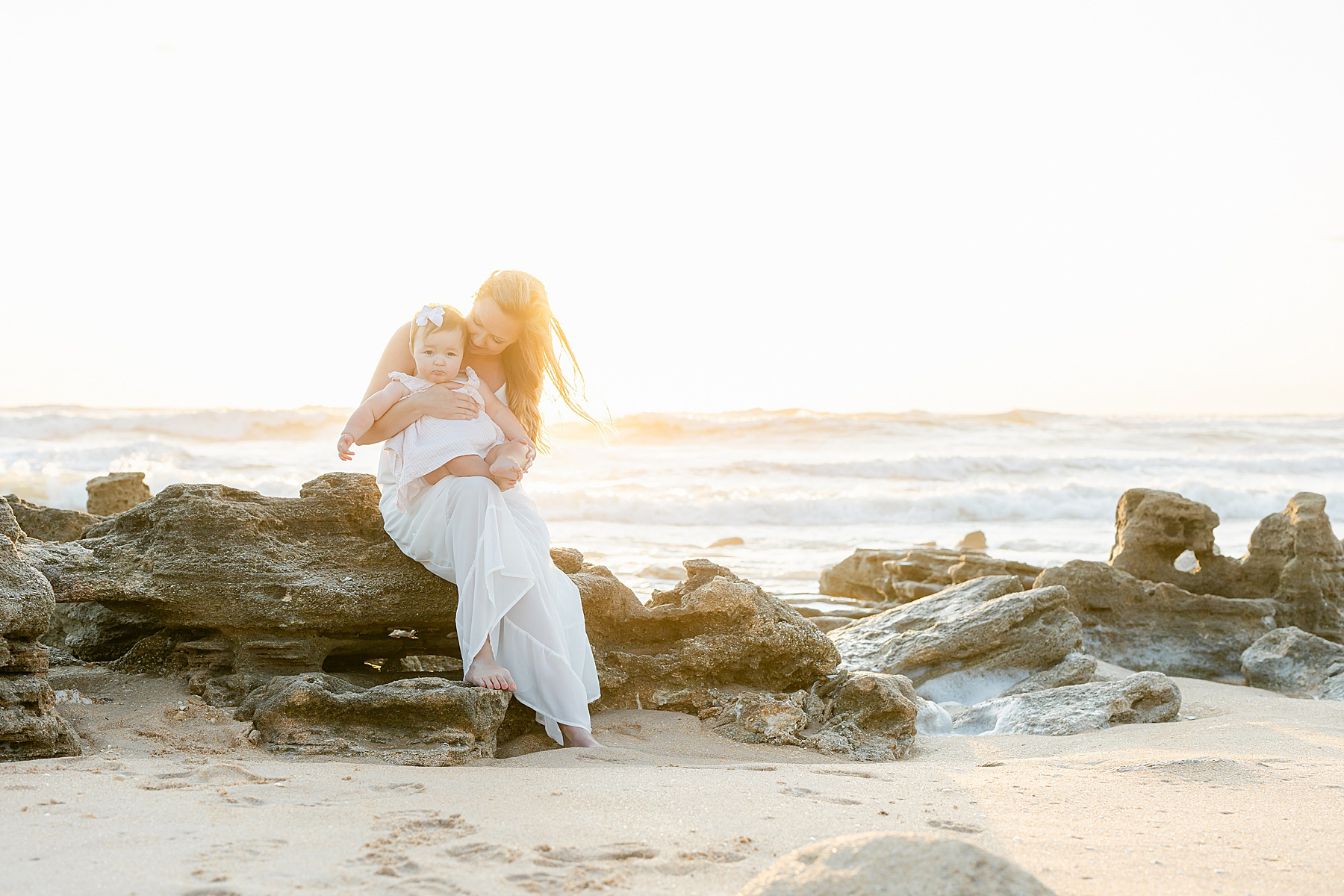 woman with long blond hair in long white dress holding baby girl on the rocks at the beach at sunrise
