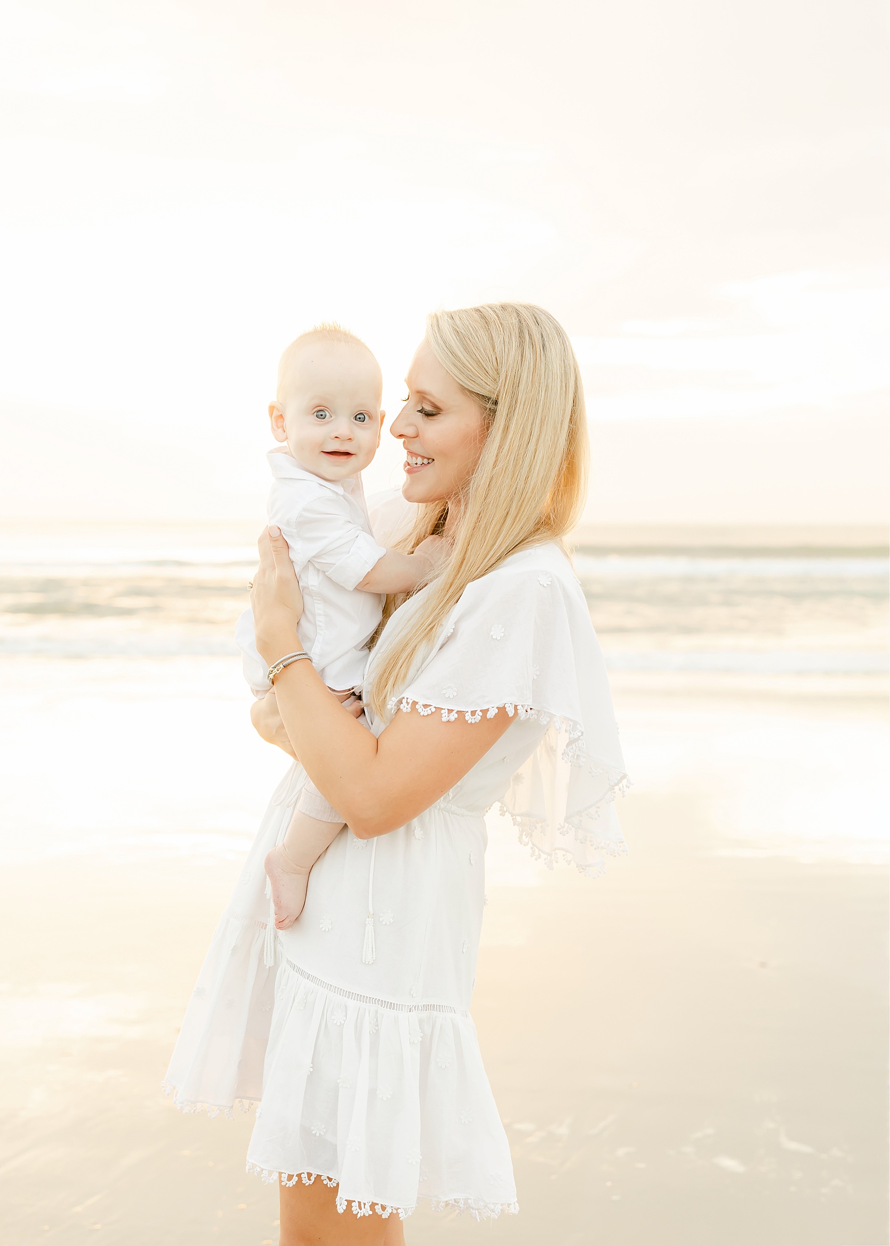 woman in white dress on the beach at sunrise holding baby boy in light and airy light