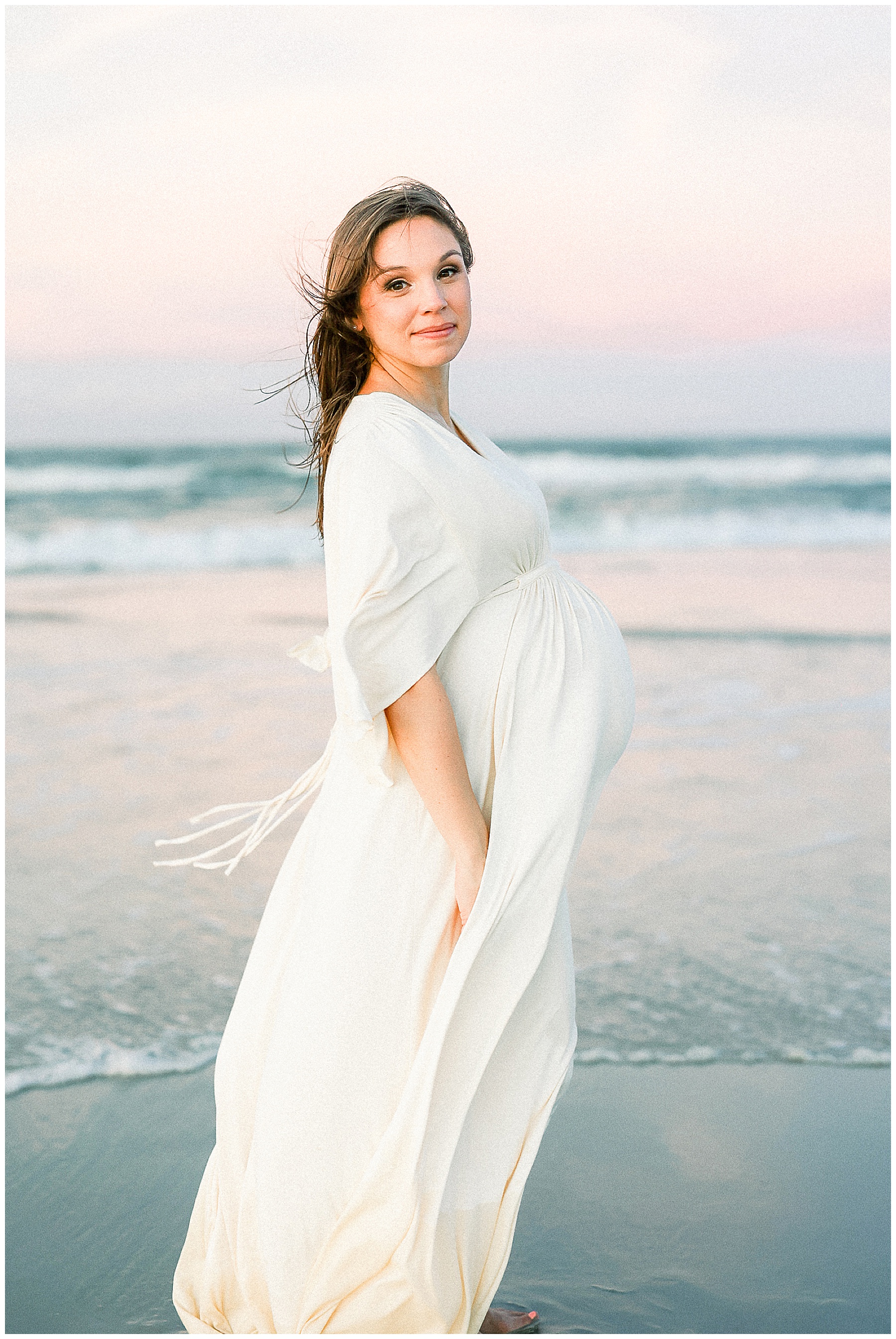 pregnant woman in cream maxi dress at sunset standing with pastel skies against the water in saint augustine beach florida