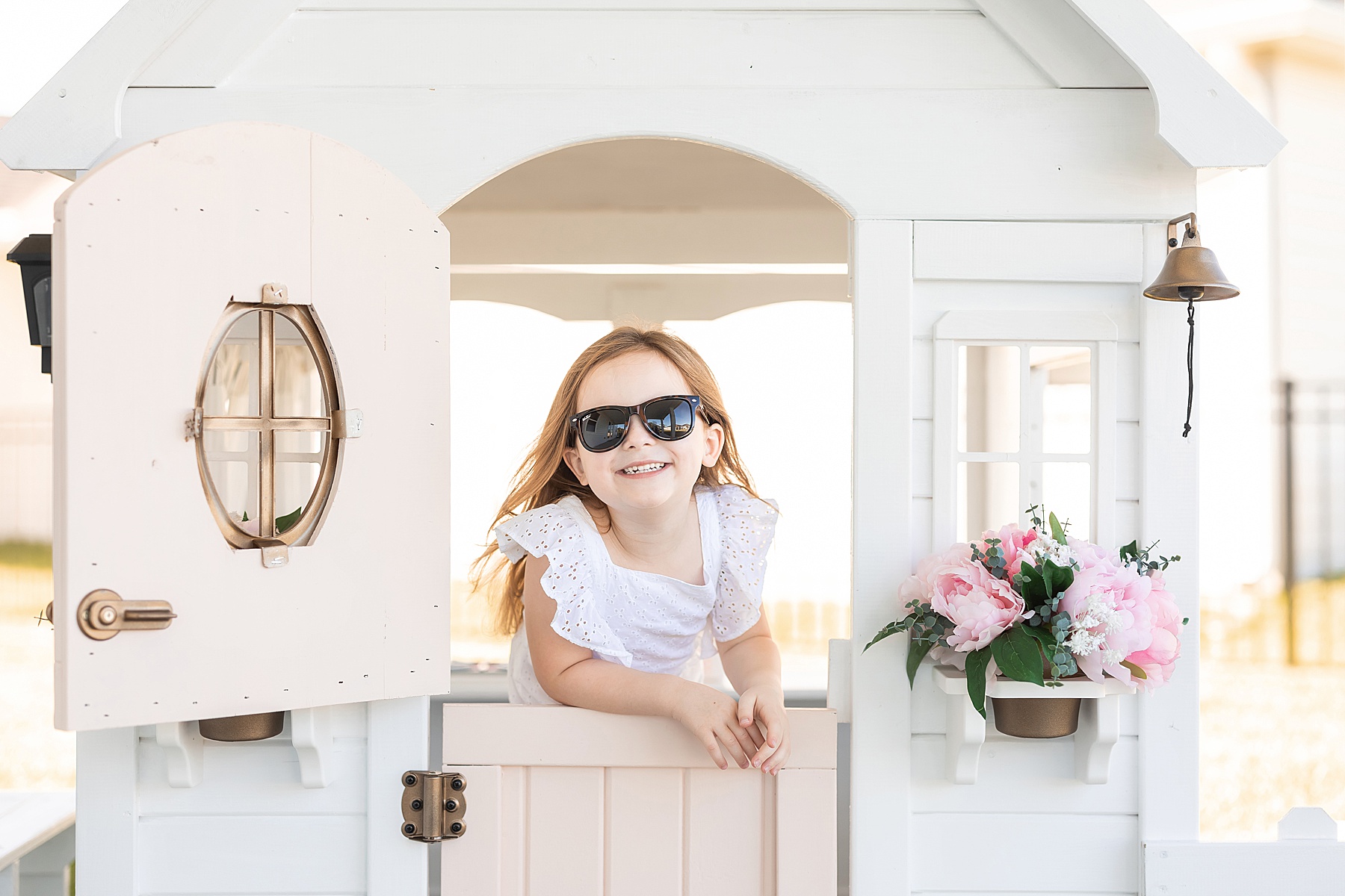 little girl in white jumpsuit standing inside of kids playhouse with pink door