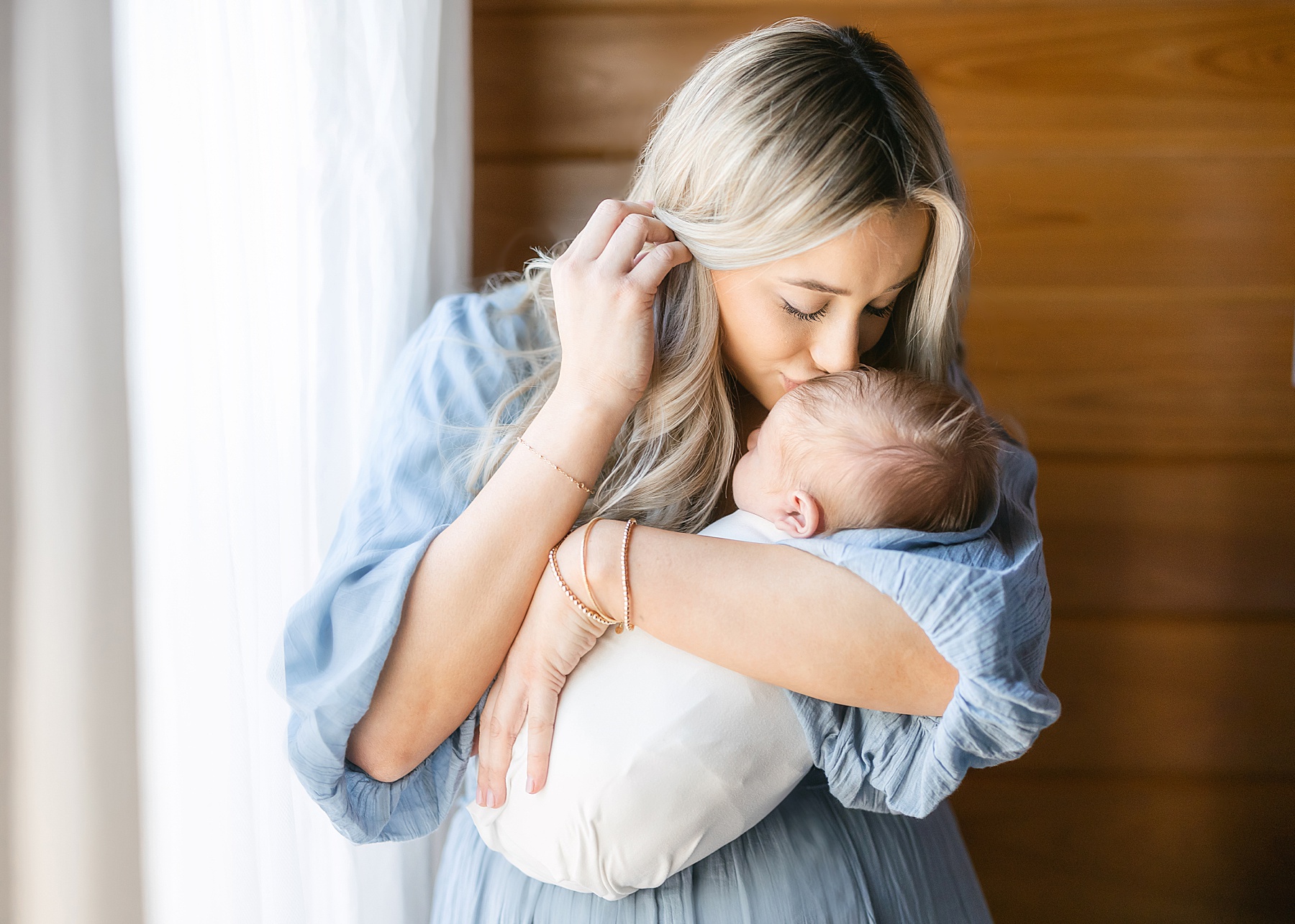 blond haired woman in long blue dress kissing newborn baby boy wrapped in white swaddle by window