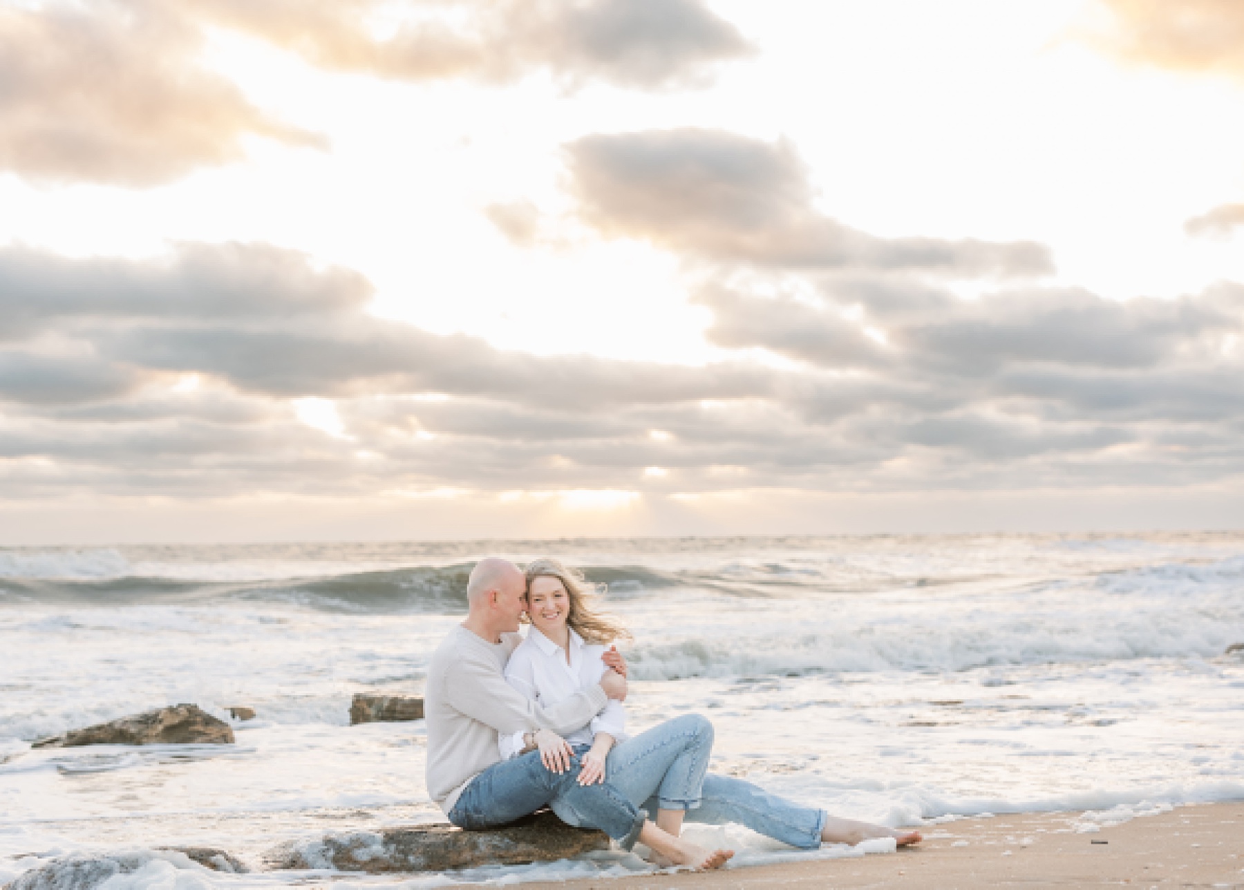 couple in jeans and white shirts sitting on a rock on the beach in st. augustine