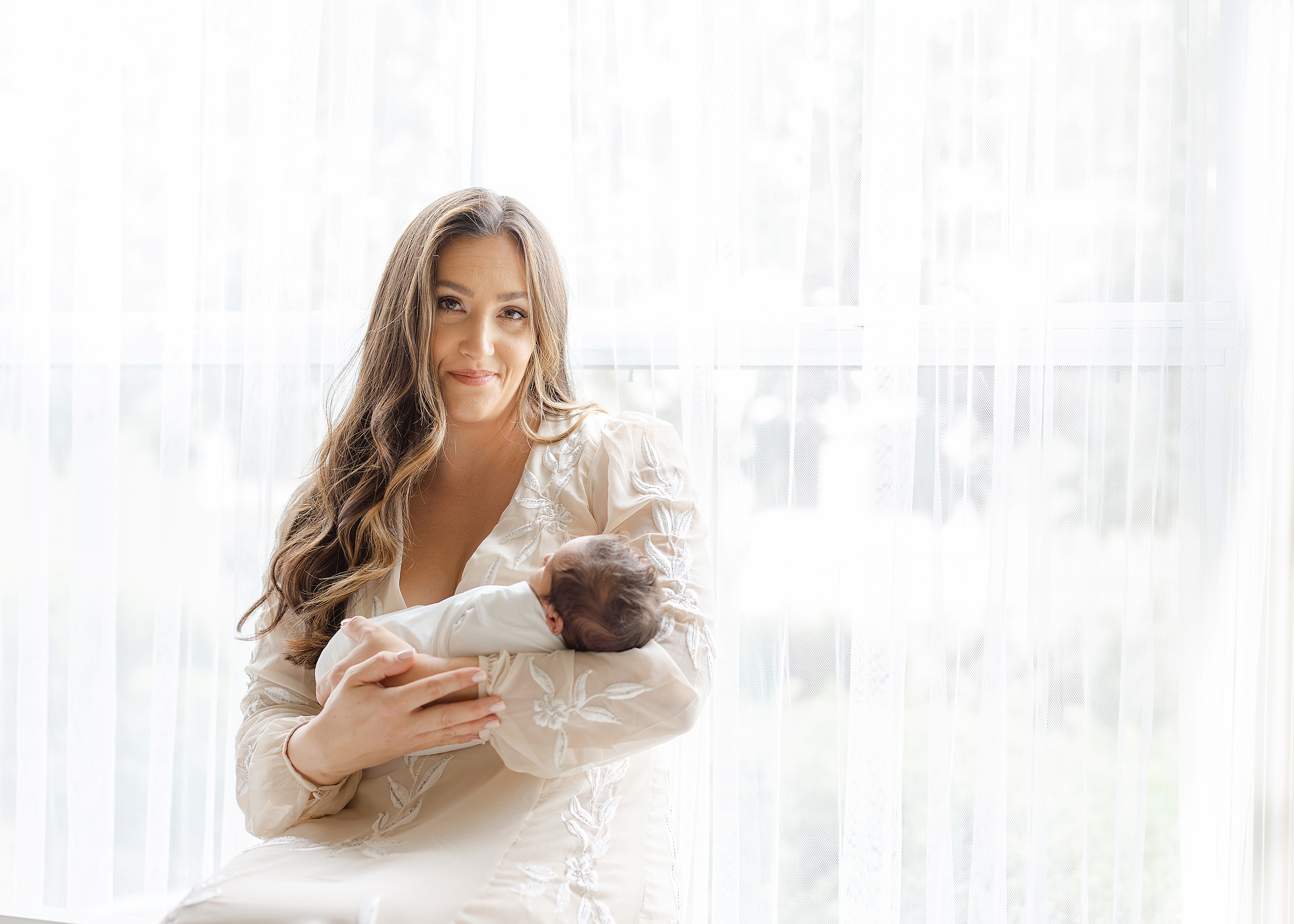 Woman holding newborn baby boy in front of window with white sheers.