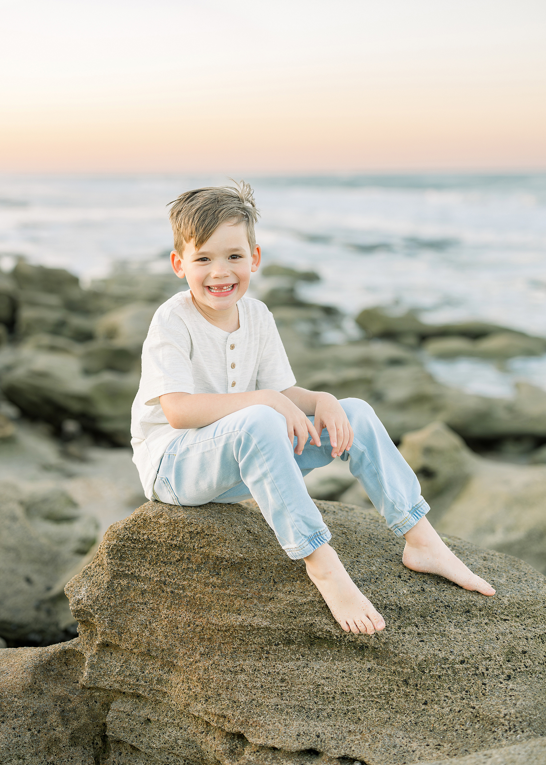 A beach portrait of a little boy in a cream shirt and jeans at sunset.