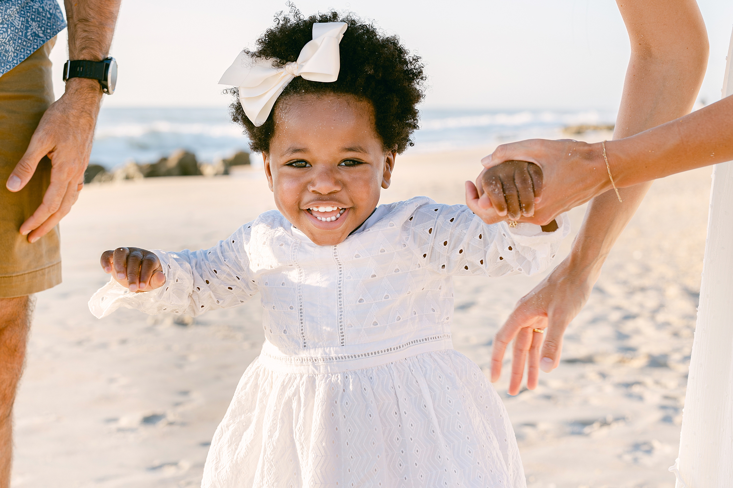 A little black girl in a white dress smiles at the camera on the beach at sunrise.